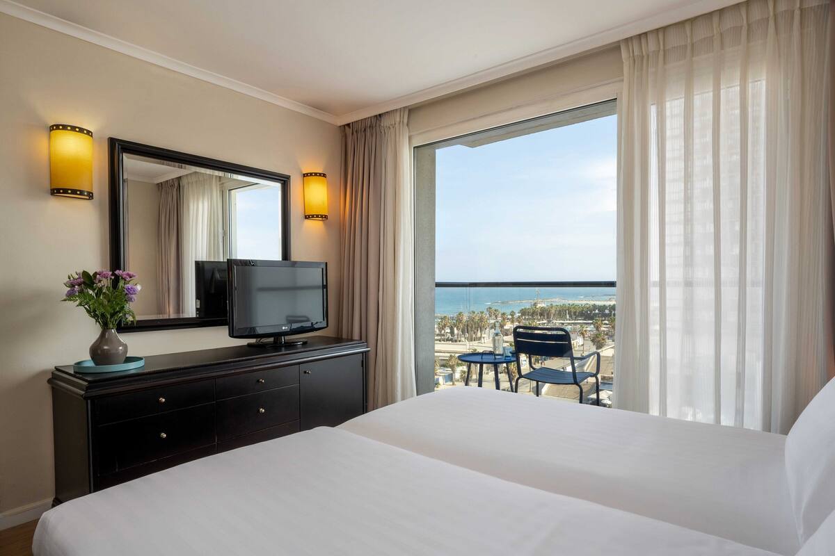 Une chambre d’hôtel avec un grand lit, un téléviseur posé sur une commode, un vase rempli de fleurs et une vue sur l’océan.