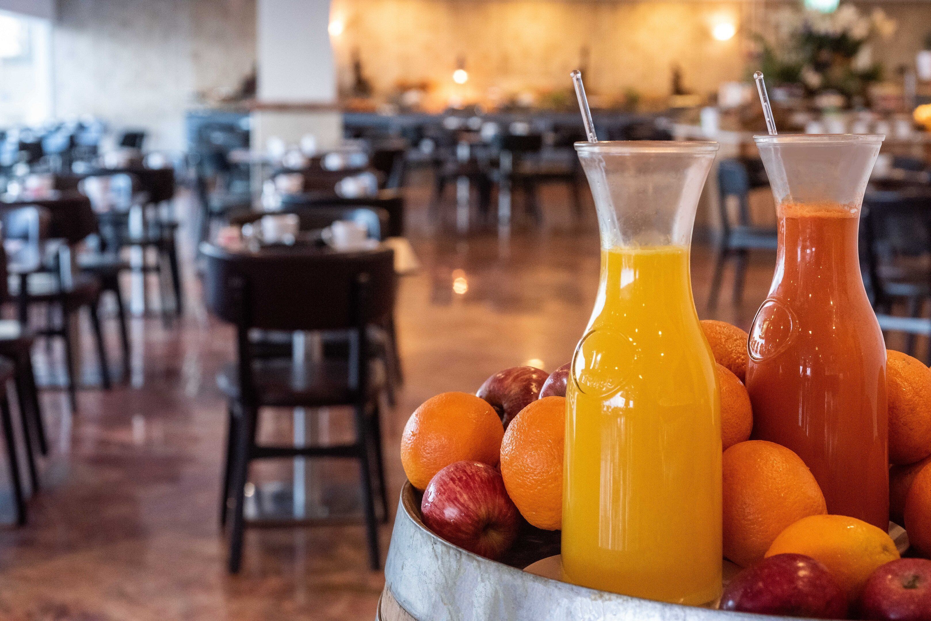 A dining area with tables and chairs, two pitchers of orange juice, and a bowl of fruits.
