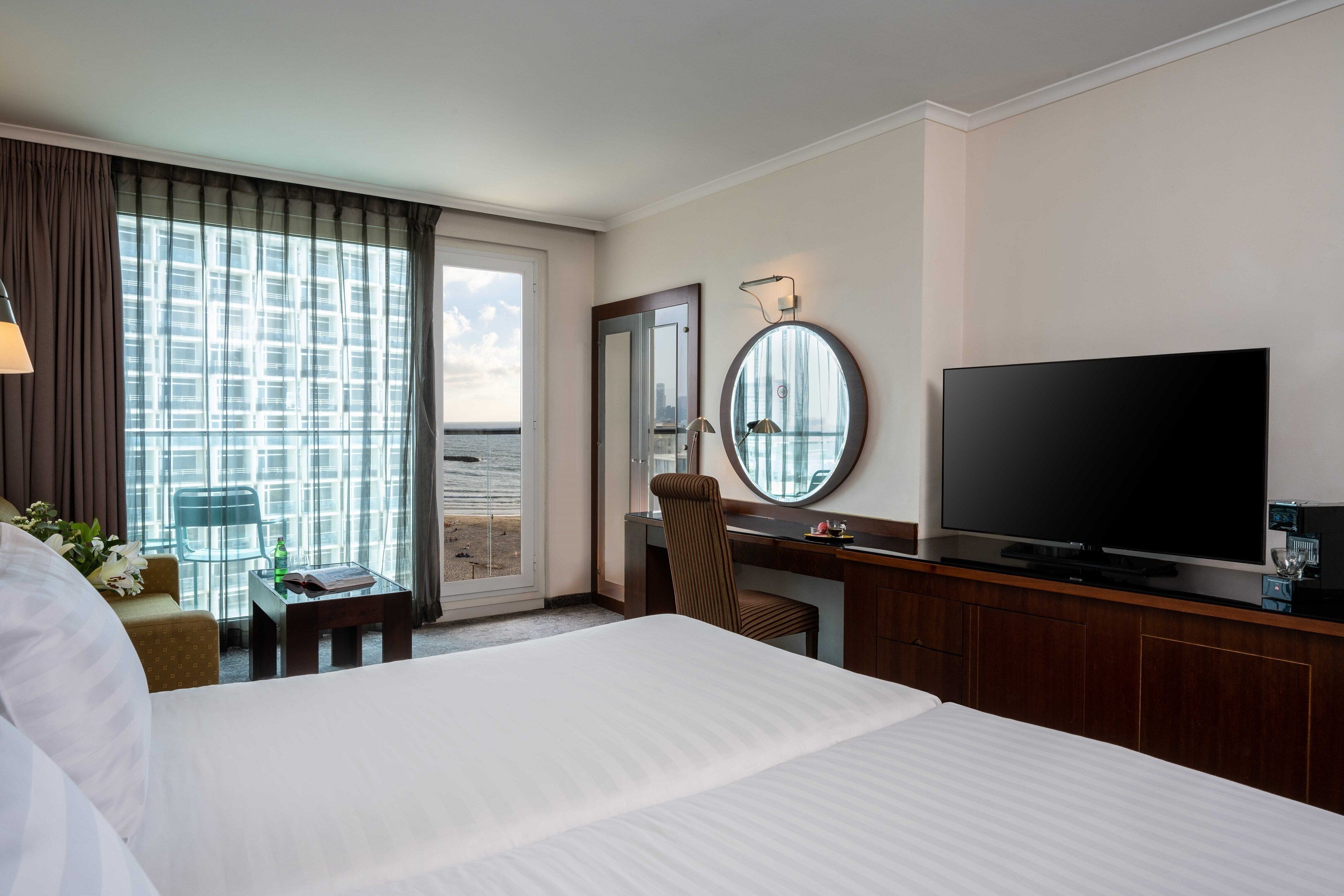 A hotel room with a large bed, a desk with a flat-screen TV, a chair, a small table with a book and a bottle, and a view of the city through large windows.