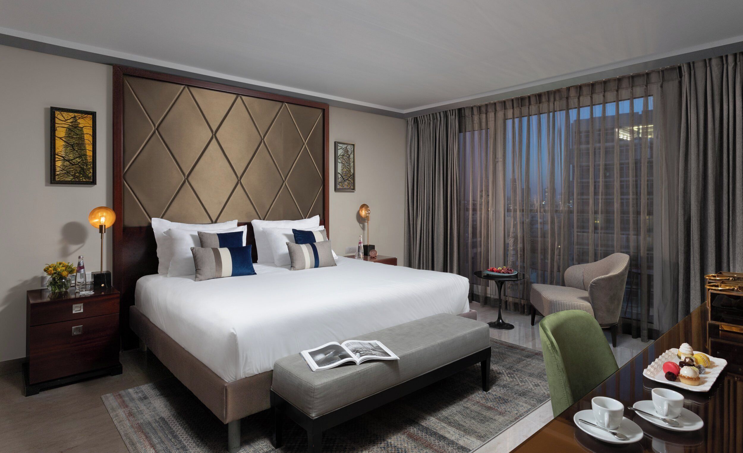 A hotel room with a large bed, a bedside table, a reading area with a chair, a small table with a tray of pastries, and a view of the city through the window.