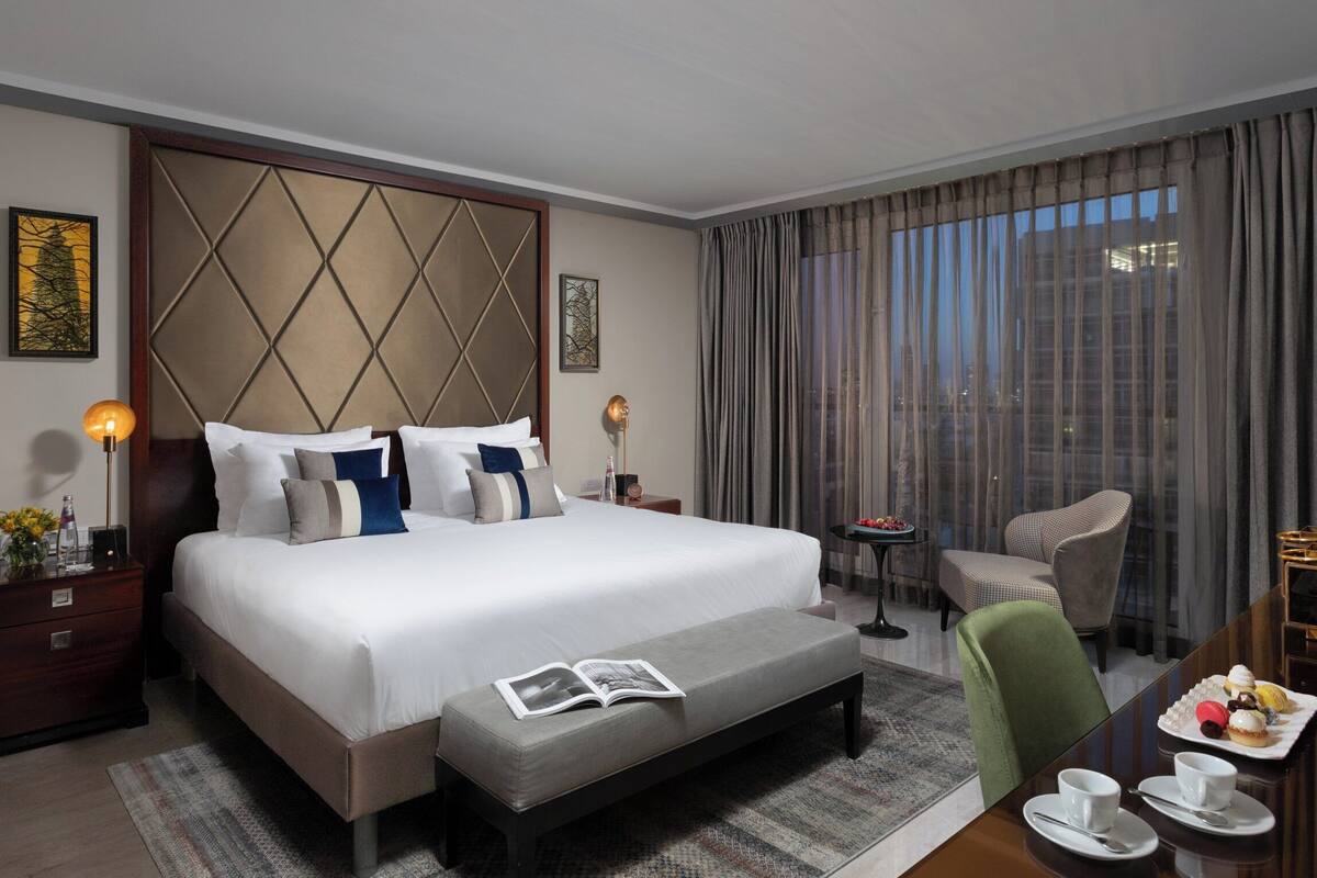 Une chambre d’hôtel avec un grand lit, une table de chevet, un coin lecture avec un fauteuil, une petite table sur laquelle se trouve un plateau de pâtisseries, et une vue sur la ville par la fenêtre.