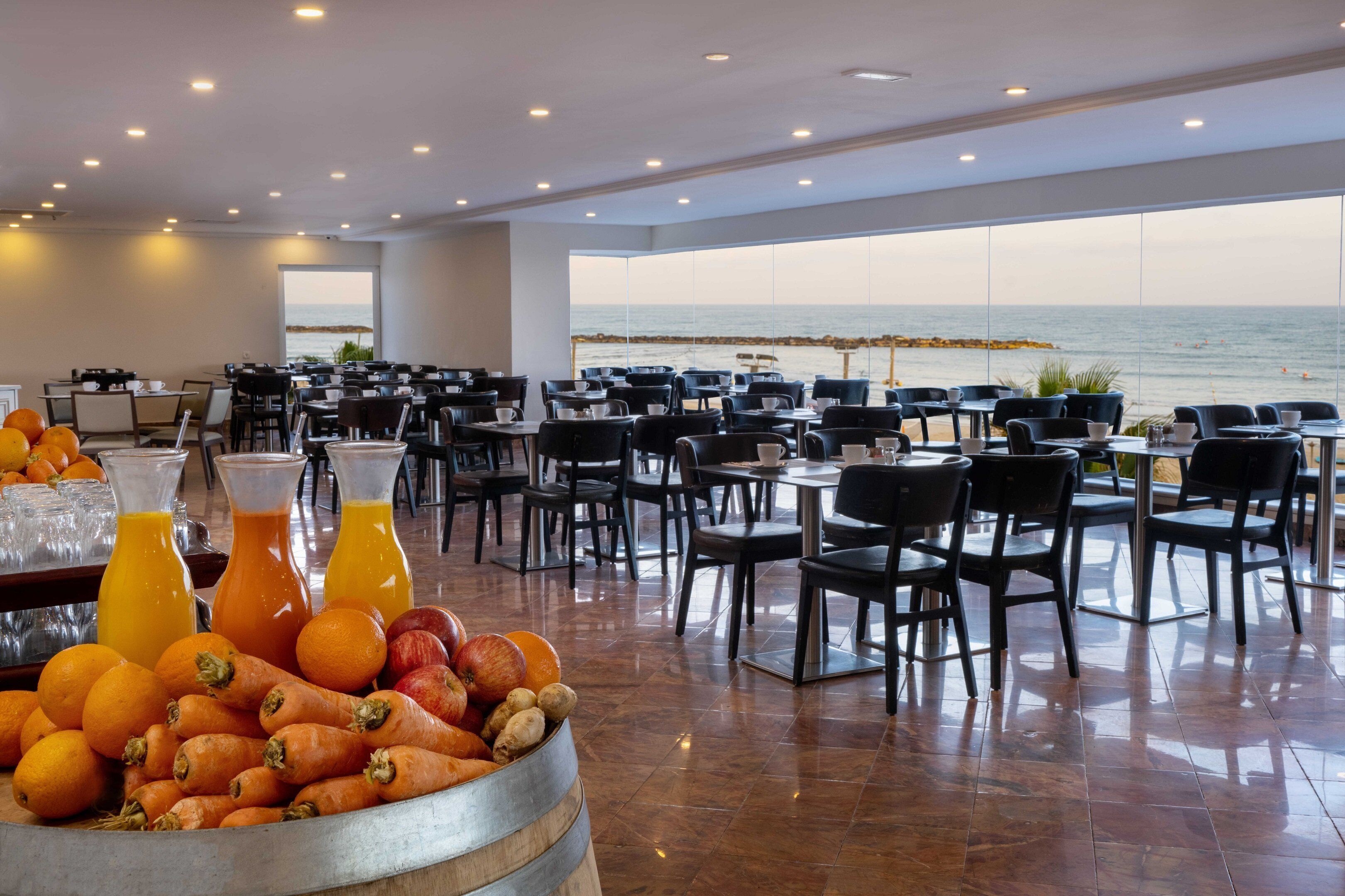 Een eetruimte met tafels en stoelen, een presentatie van fruit en sap, en uitzicht op de zee.