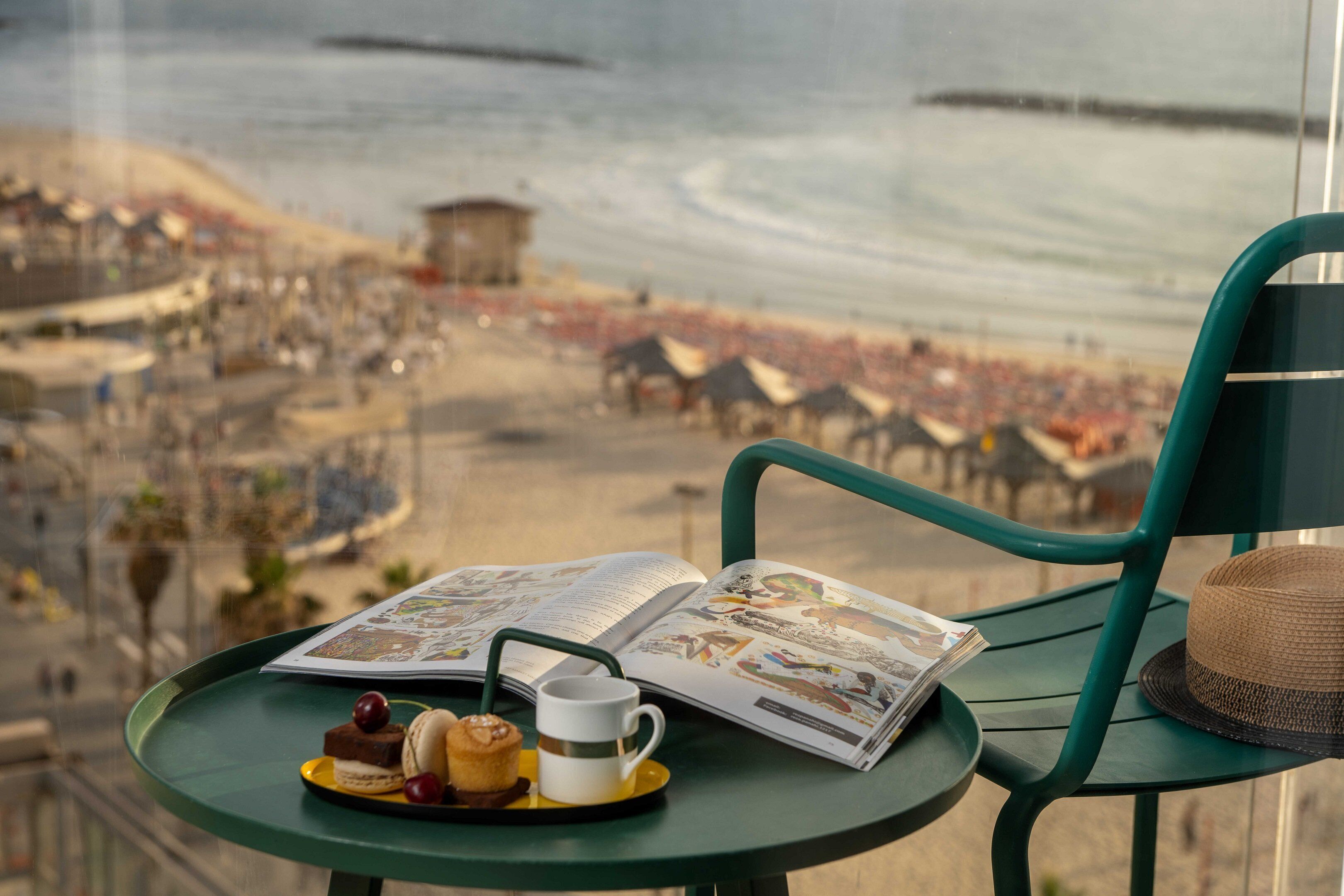 Ein Tisch mit einem Buch, einer Tasse und Gebäck, mit Blick auf einen Strand mit Menschenmassen und Gebäuden.