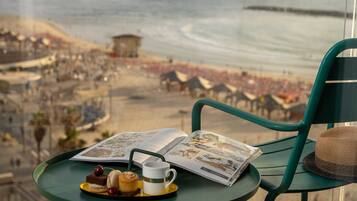 Ein Tisch mit einem Buch, einer Tasse und Gebäck, mit Blick auf einen Strand mit Menschenmassen und Gebäuden.