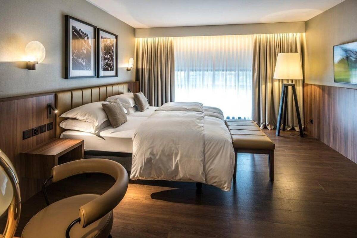 A hotel room with a large bed, a bedside table, a chair, a floor lamp, and a view of the outdoors through the window.