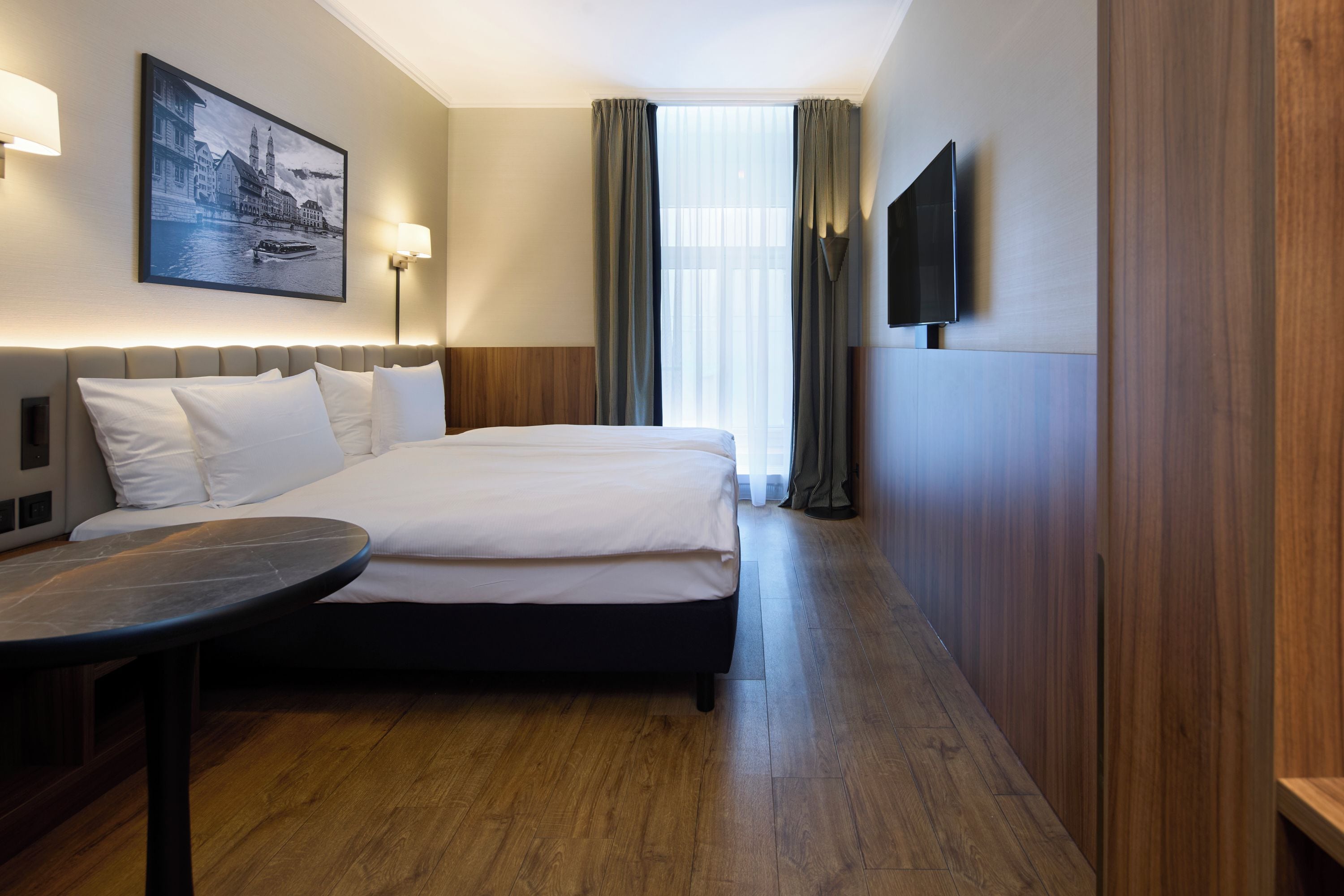 A hotel room with a bed, a desk, a TV, and a painting on the wall.