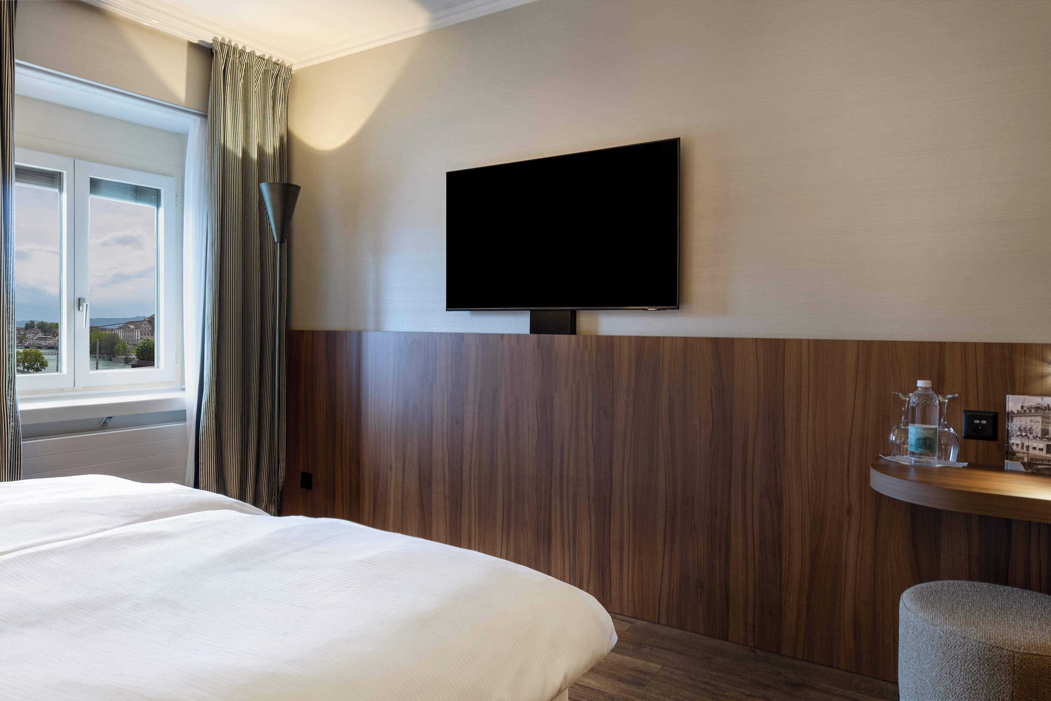 Hotellihuone, jossa on sänky, seinälle asennettu taulutelevisio, puupanelointi ja ikkuna, jossa on verhot.