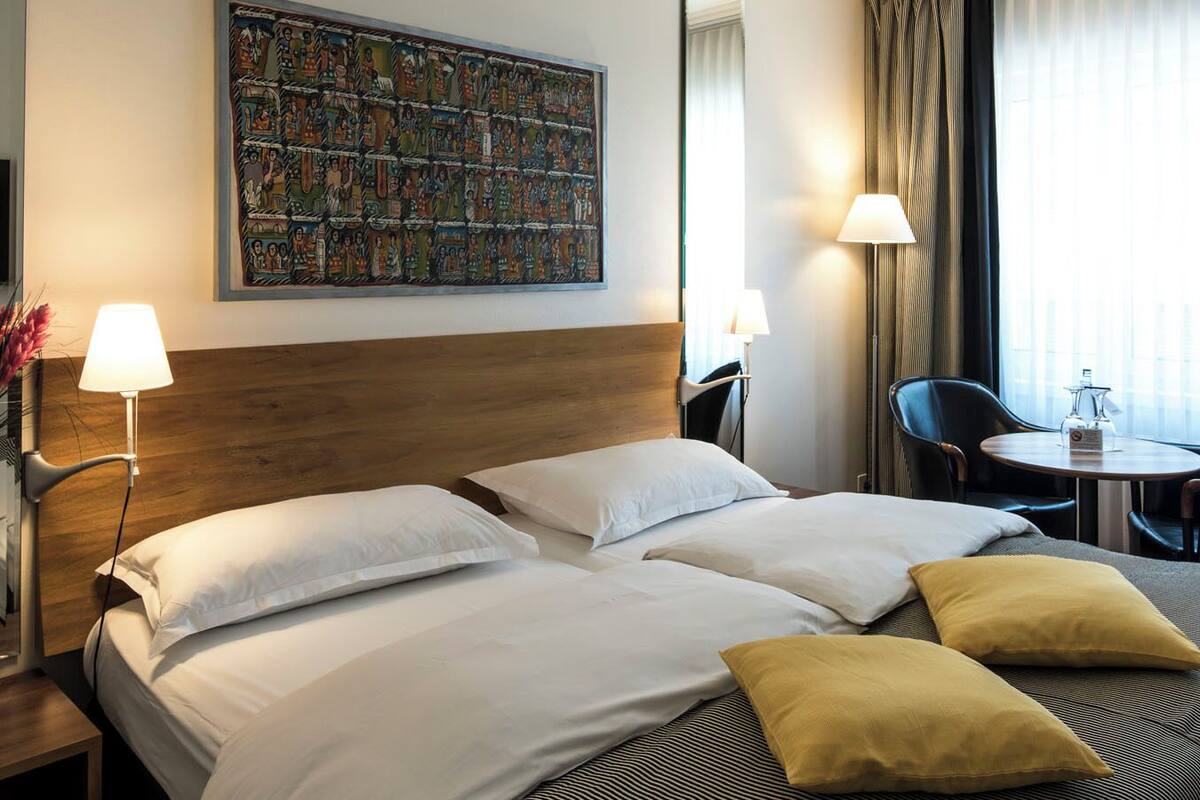 A hotel room with a large bed, two bedside tables, a small round table with chairs, a painting on the wall, and a window with curtains.
