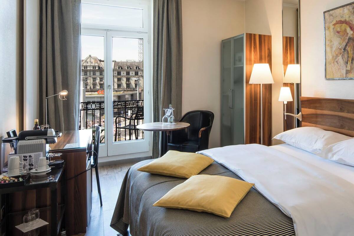 A hotel room with a bed, bedside lamps, a desk with a chair, a small table, and a window with curtains.