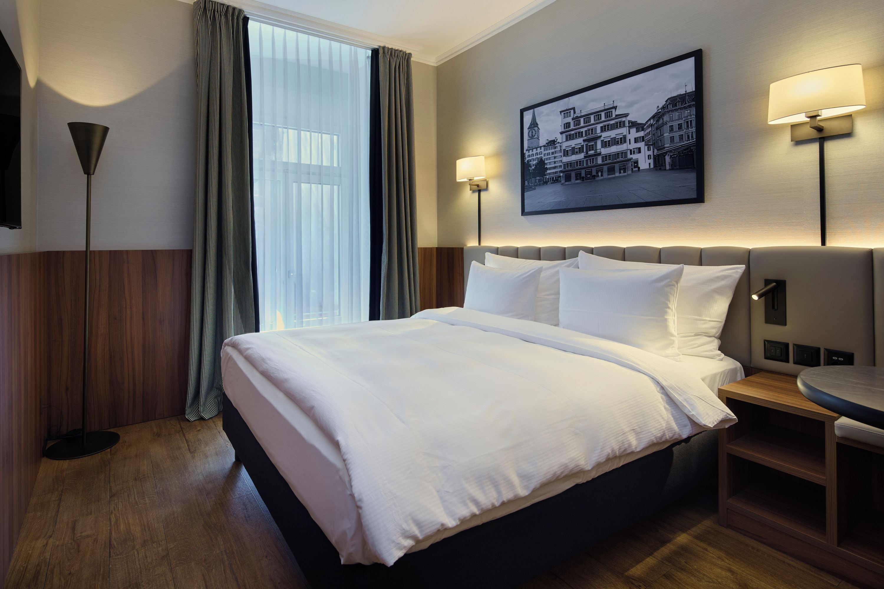 Ein Hotelzimmer mit einem großen Bett, einem Nachttisch, einem runden Tisch, einer Wandlampe und einem gerahmten Bild.