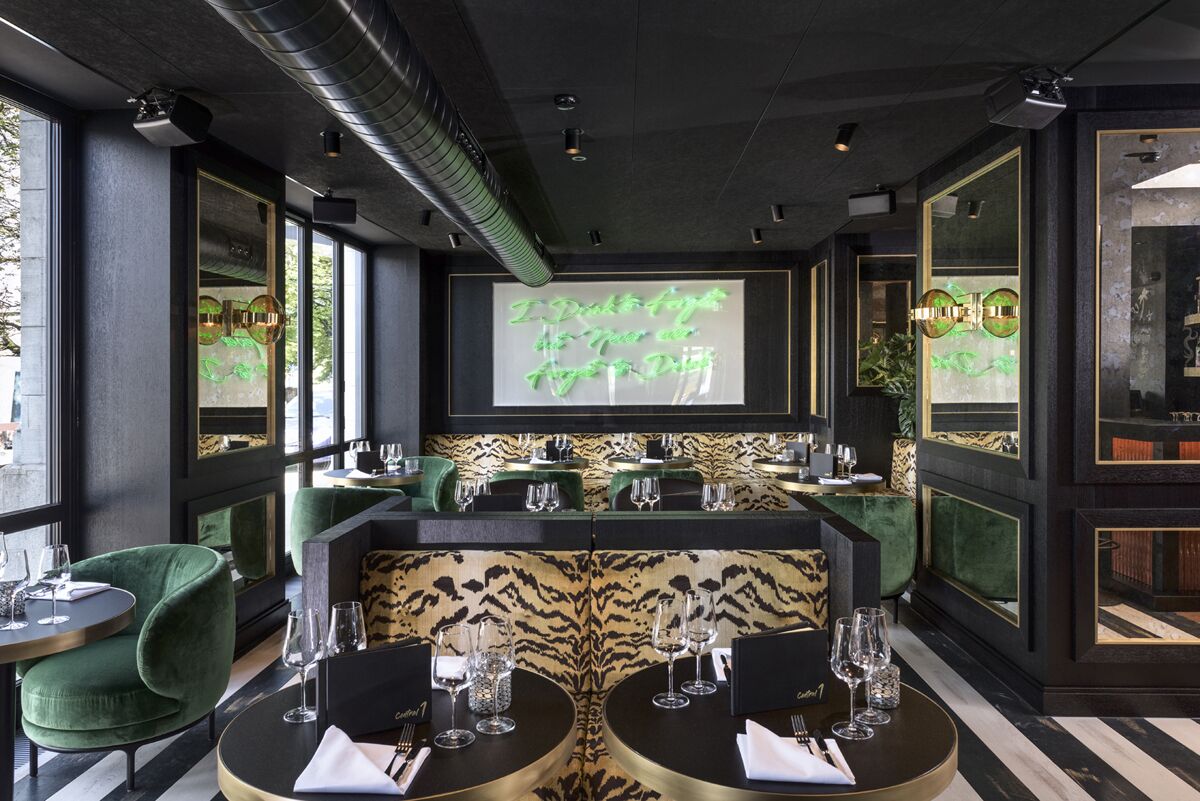 A modern restaurant interior with a central bar, green upholstered chairs, and a patterned wall.