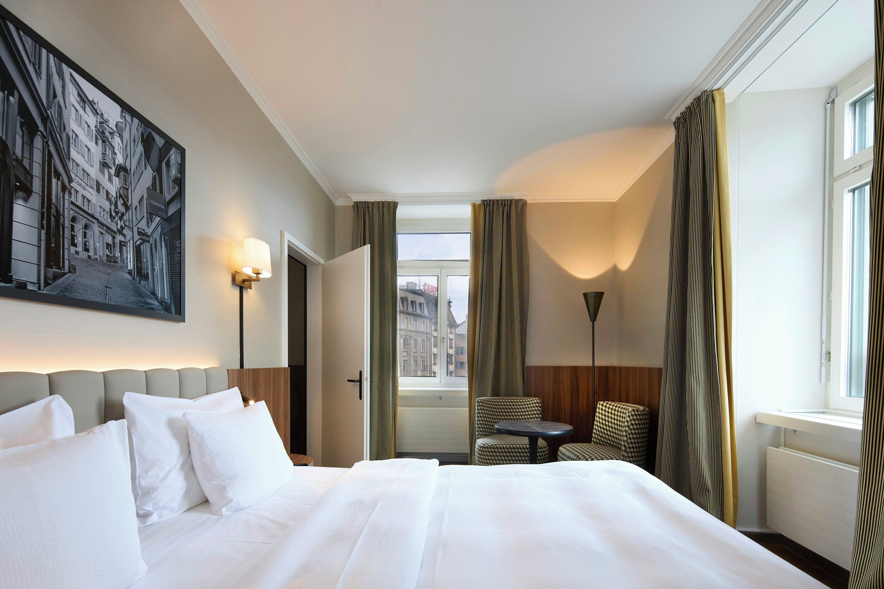 A hotel room with a large bed, a chair, a table, a painting on the wall, and a view of buildings outside the window.