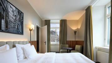 A hotel room with a large bed, a chair, a table, a painting on the wall, and a view of buildings outside the window.
