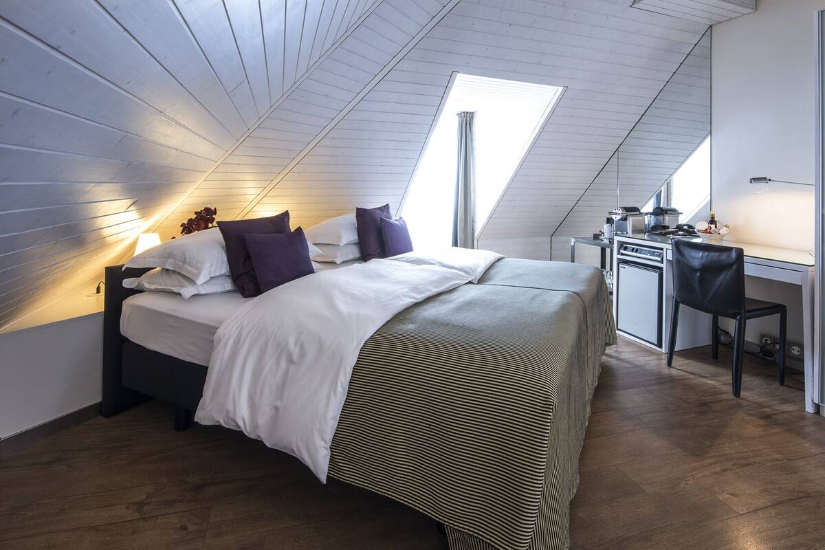A modern loft bedroom with a sloped ceiling, a bed with white and striped bedding, a desk with a chair, and a skylight.
