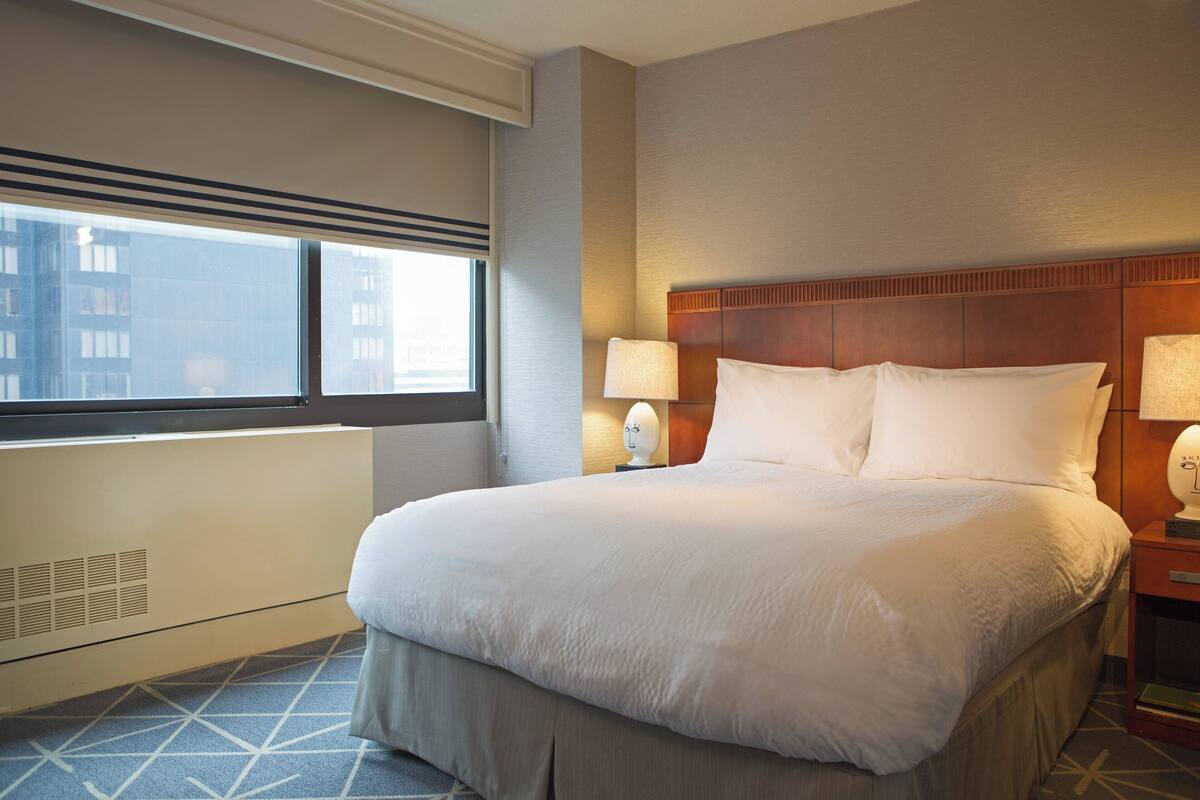 A hotel room with a large bed, two bedside lamps, a radiator, and a window with a view of buildings.