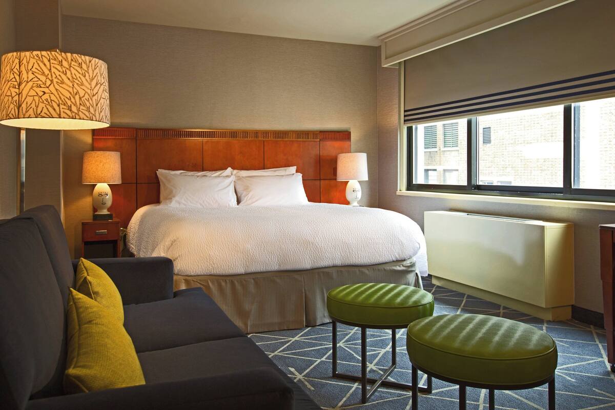 A hotel room with a large bed, two bedside lamps, a sofa, two green ottomans, and a window with a view of buildings.