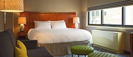 A hotel room with a large bed, two bedside lamps, a sofa, two green ottomans, and a window with a view of buildings.
