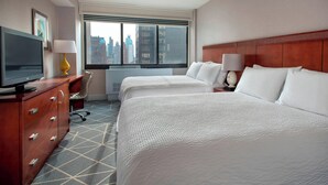 A hotel room with two beds, a TV, a desk, and a window with a city view.