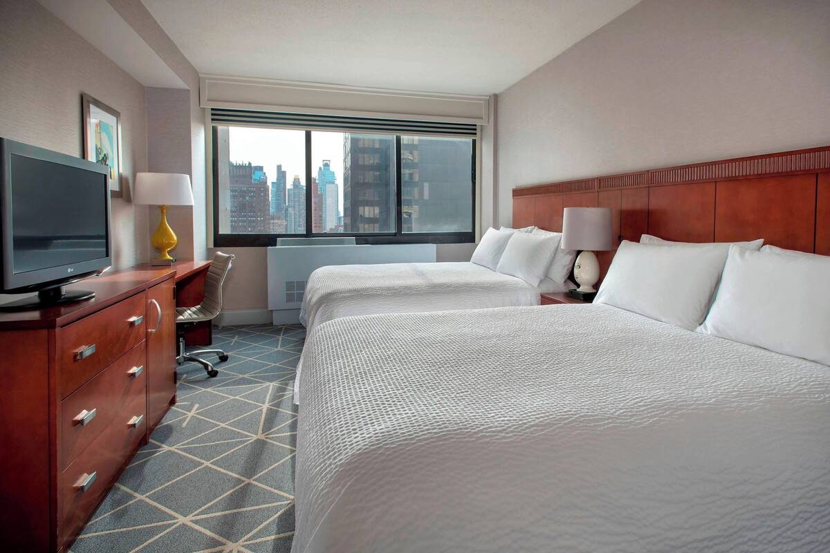 A hotel room with two beds, a TV, a desk, and a window with a city view.
