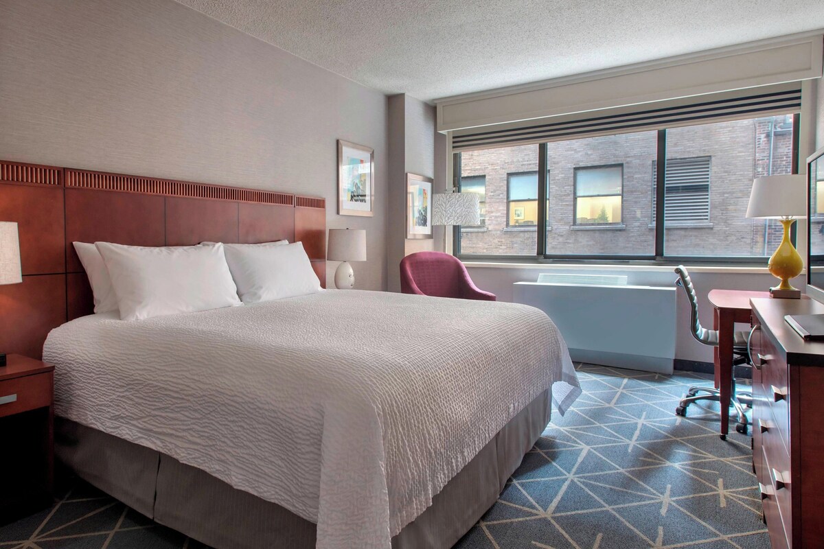 A hotel room with a large bed, a desk with a lamp, a chair, and a window with a view of buildings.