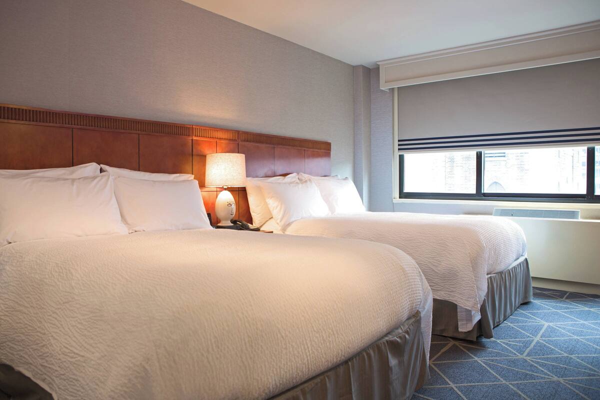 A hotel room with two beds, a headboard, a nightstand with a lamp, and a window with blinds.
