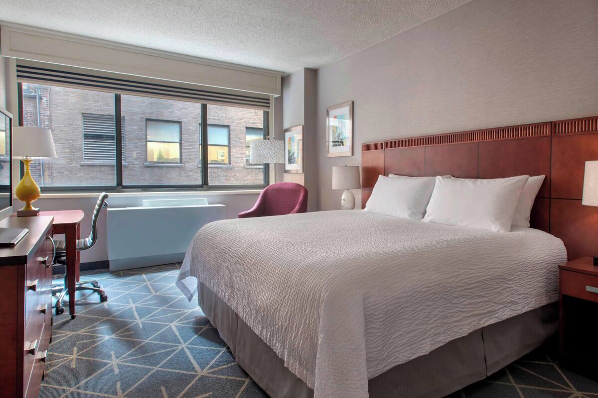 A hotel room with a large bed, a desk with a lamp, a chair, and a window with blinds.