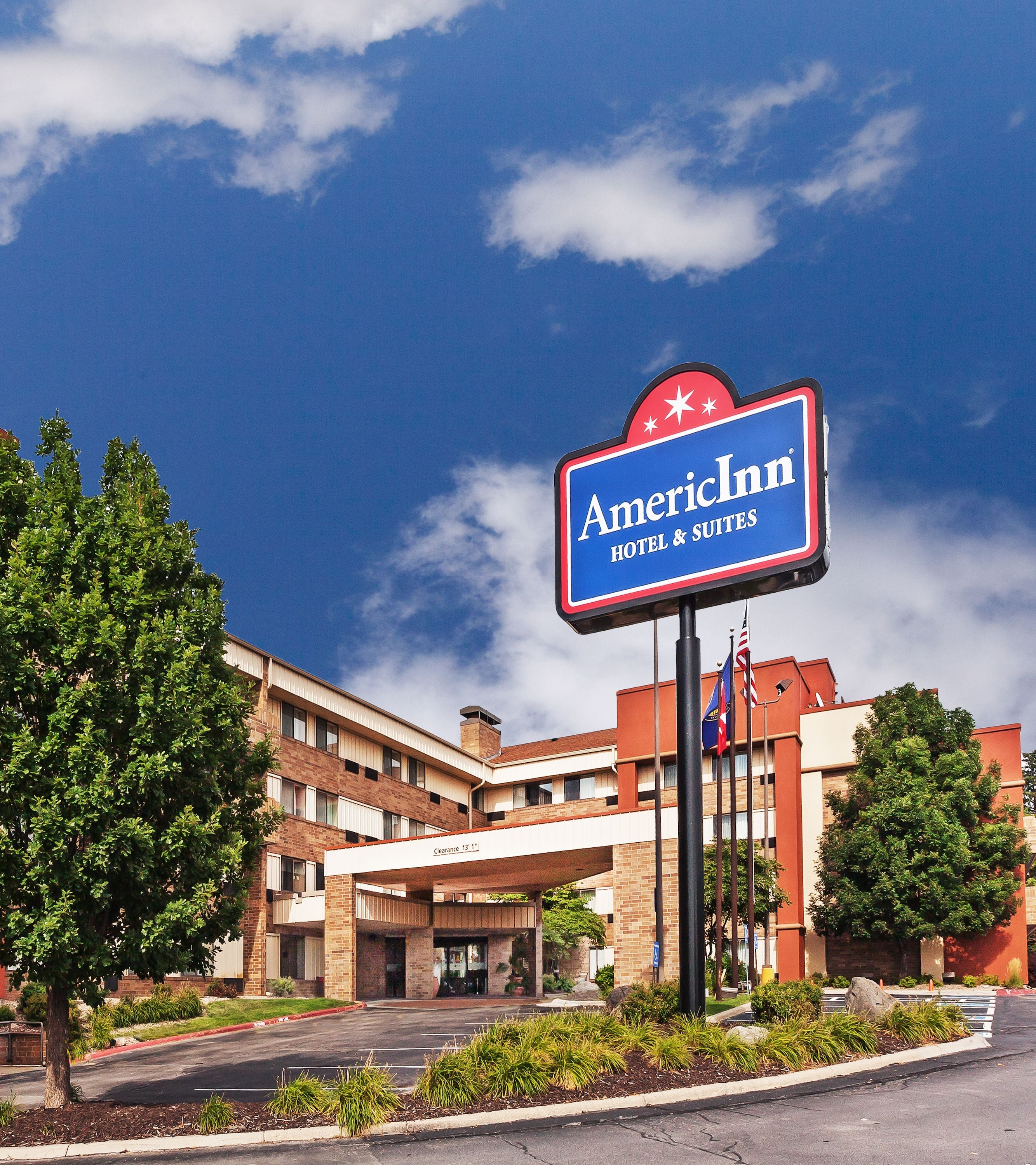 An American Inn hotel sign with a clear view of the building's entrance, a parking lot, and surrounding greenery.