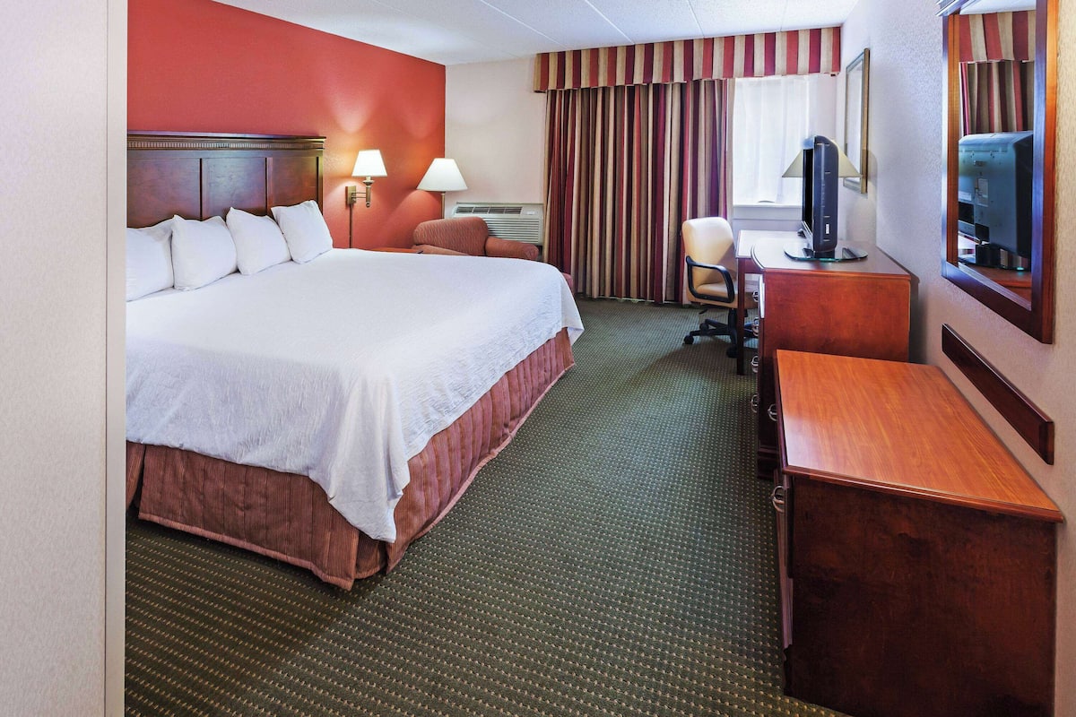 Ein Hotelzimmer mit einem großen Bett, einem Schreibtisch mit Computer, einem Sessel und einem Fernseher.