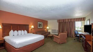 A hotel room with a large bed, a sofa, a desk with a chair, a television, and an air conditioning unit. - AmericInn by Wyndham Omaha (Omaha)