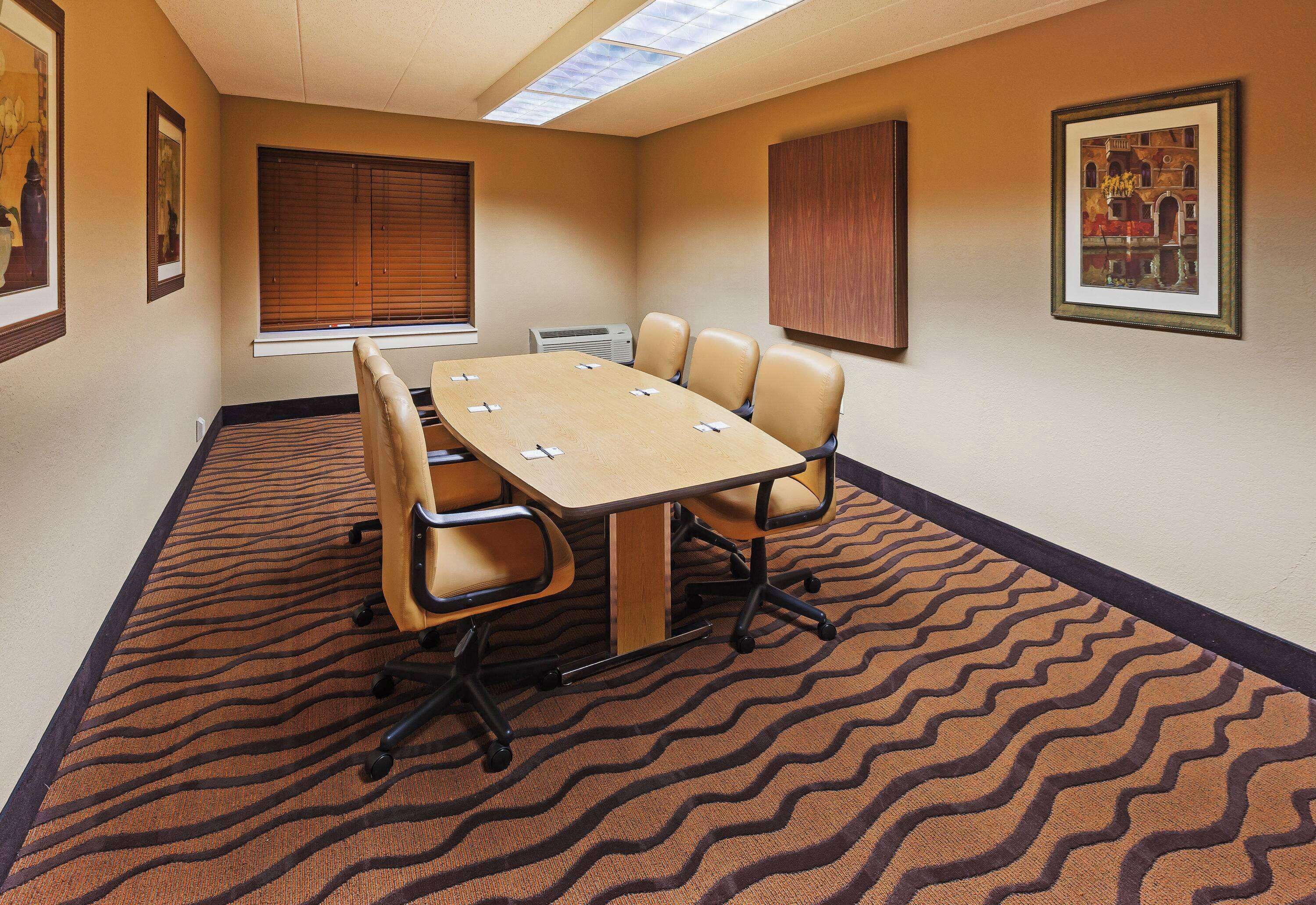 A conference room with a large table, four chairs, a window with blinds, and a patterned carpet.