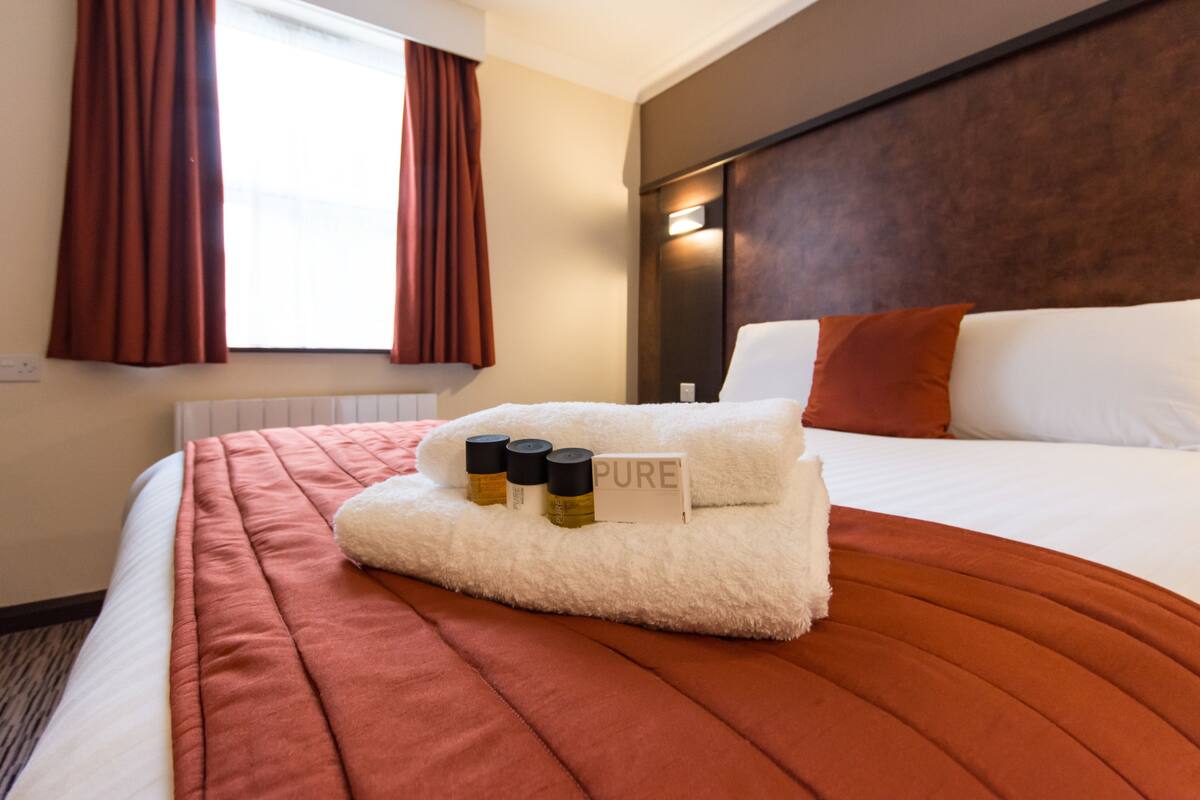 A hotel room with a neatly made bed, a window with curtains, a headboard, and a sign on the bed.