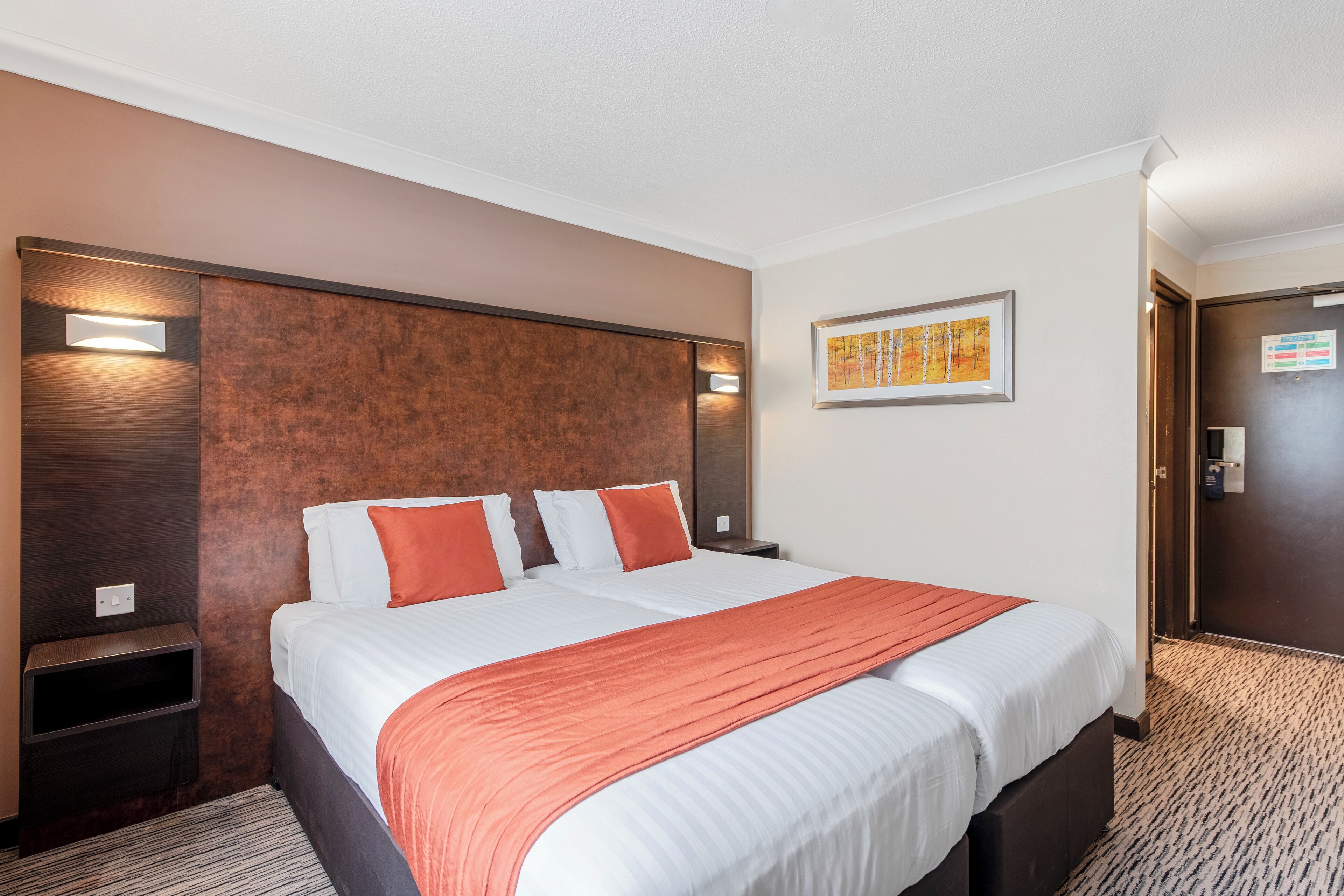 A hotel room with a large bed, two bedside tables, a headboard, a framed picture on the wall, and a door.