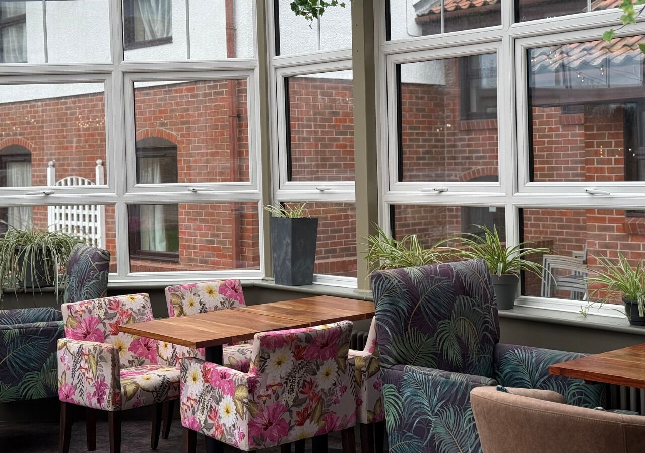 A cozy seating area with floral-patterned chairs and a wooden table, large windows with a view of a brick building, and potted plants on the windowsill.
