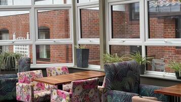 A cozy seating area with floral-patterned chairs and a wooden table, large windows with a view of a brick building, and potted plants on the windowsill.