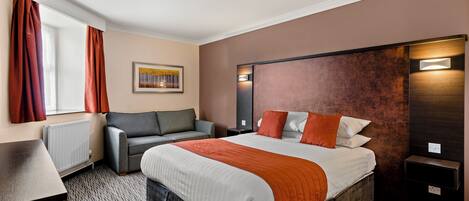 A hotel room with a large bed, a gray armchair, a small table, and a window with curtains.