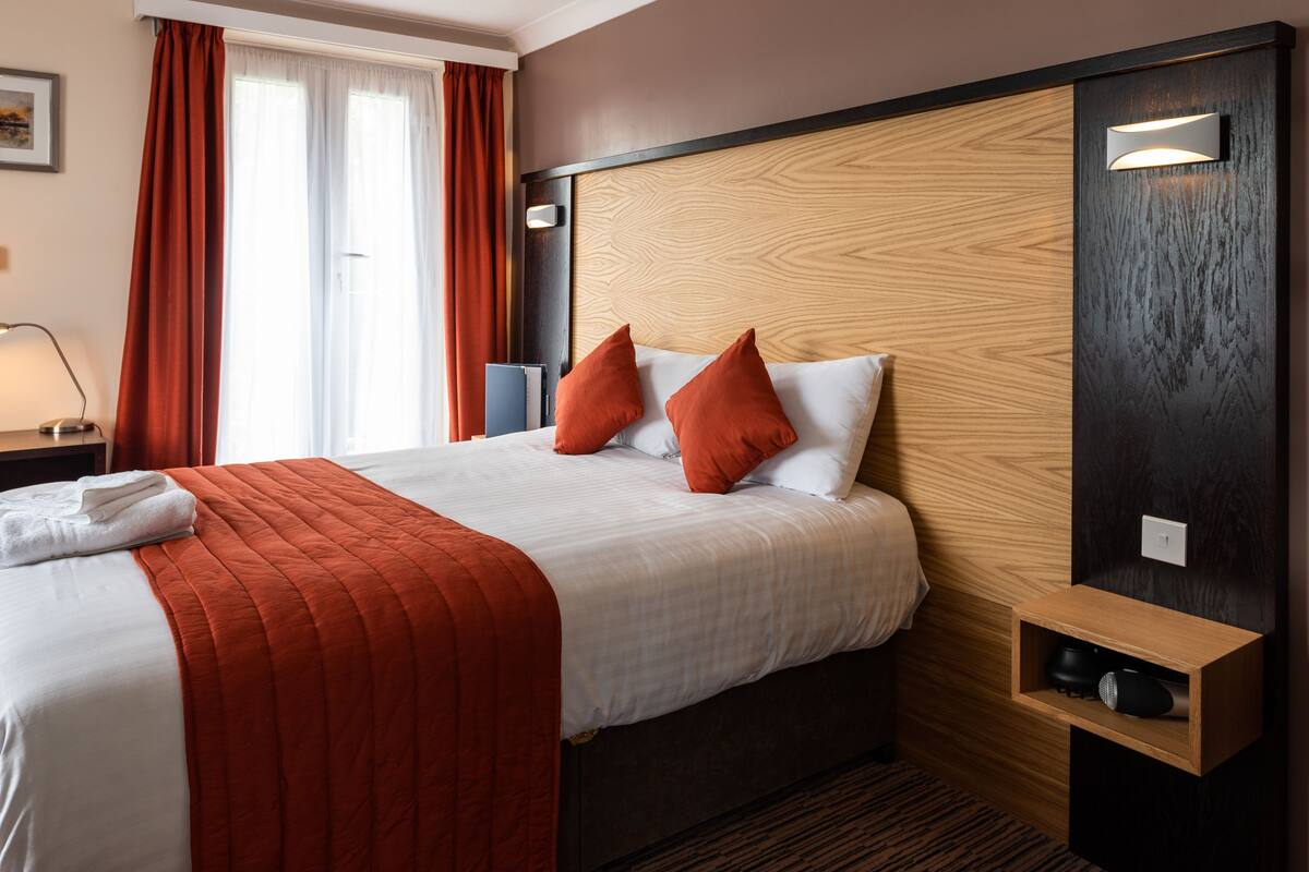 A hotel room with a large bed, red and white bedding, a wooden headboard, a bedside table with a lamp, and a small shelf with a speaker.