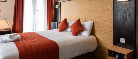 A hotel room with a large bed, red and white bedding, a wooden headboard, a bedside table with a lamp, and a small shelf with a speaker.