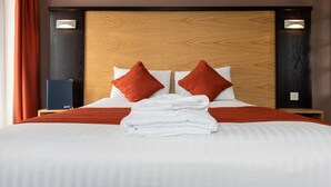 A neatly made bed with a wooden headboard, two red pillows, and a stack of white towels. - Dragonfly Hotel Bury St Edmunds (Bury St Edmunds)