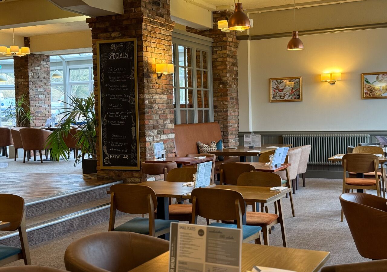 Ein Restaurant mit Backsteinwänden, Holztischen und -stühlen. An der Wand hängt eine Kreidetafel mit Tagesangeboten und Hauptspeisen.