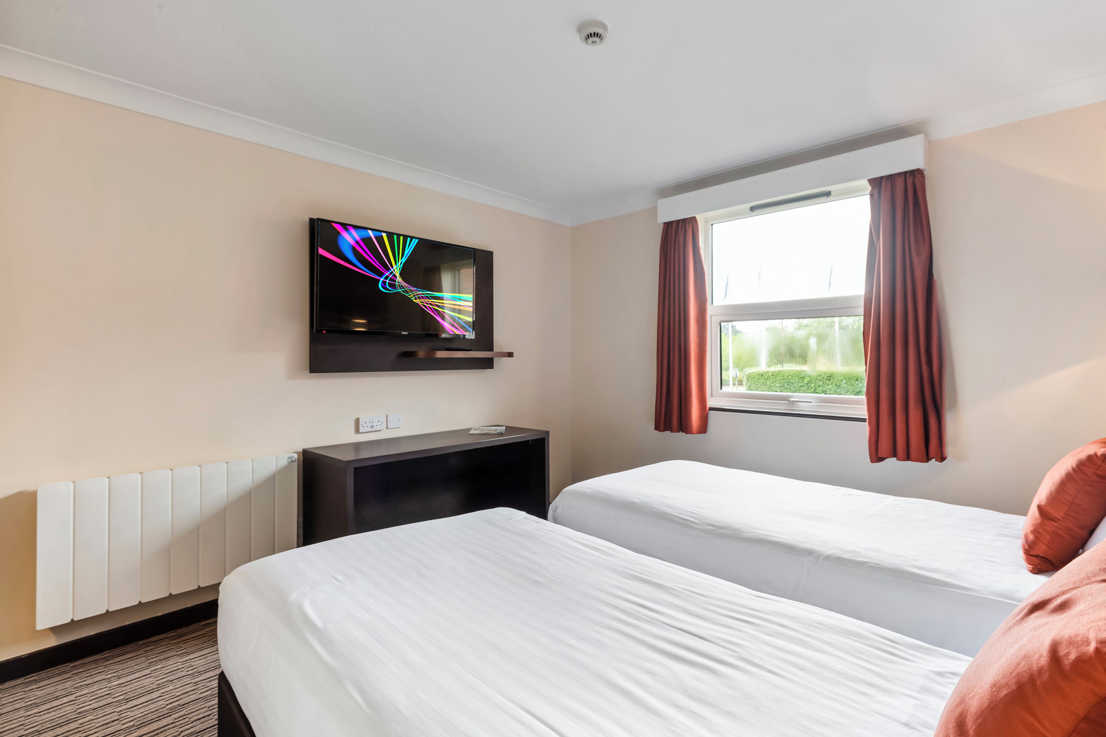 A hotel room with two beds, a TV mounted on the wall, a window with curtains, and a radiator.