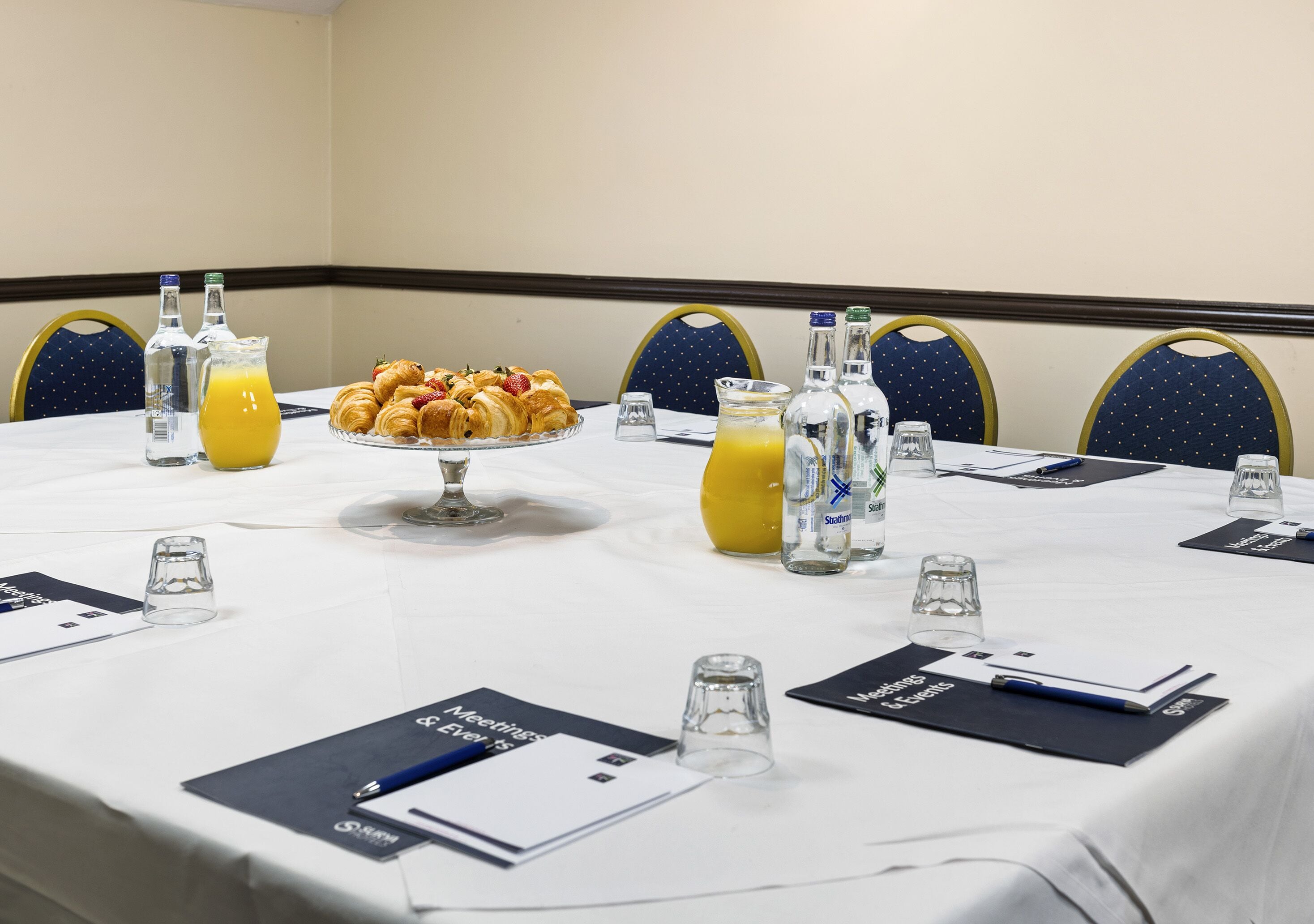 A conference table set with a fruit platter, juice, and water bottles, surrounded by chairs with name cards.