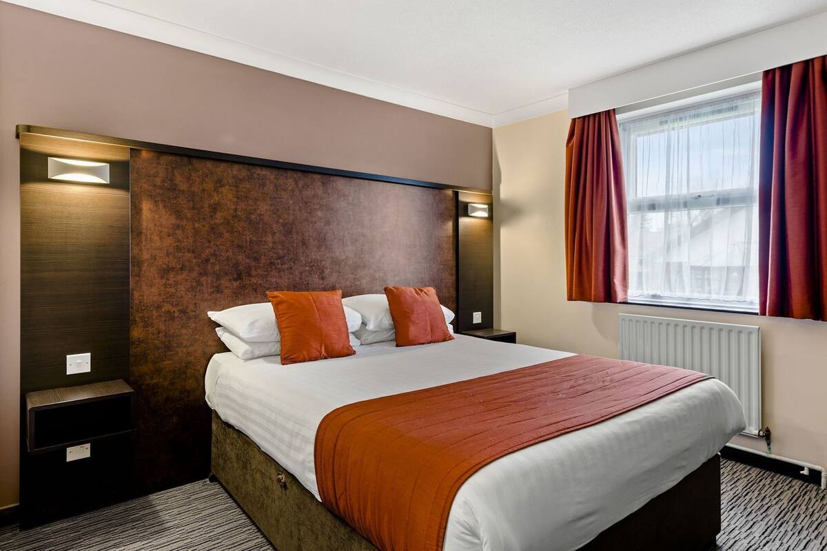 A hotel room with a bed, nightstands, a window with curtains, and a wall-mounted light.