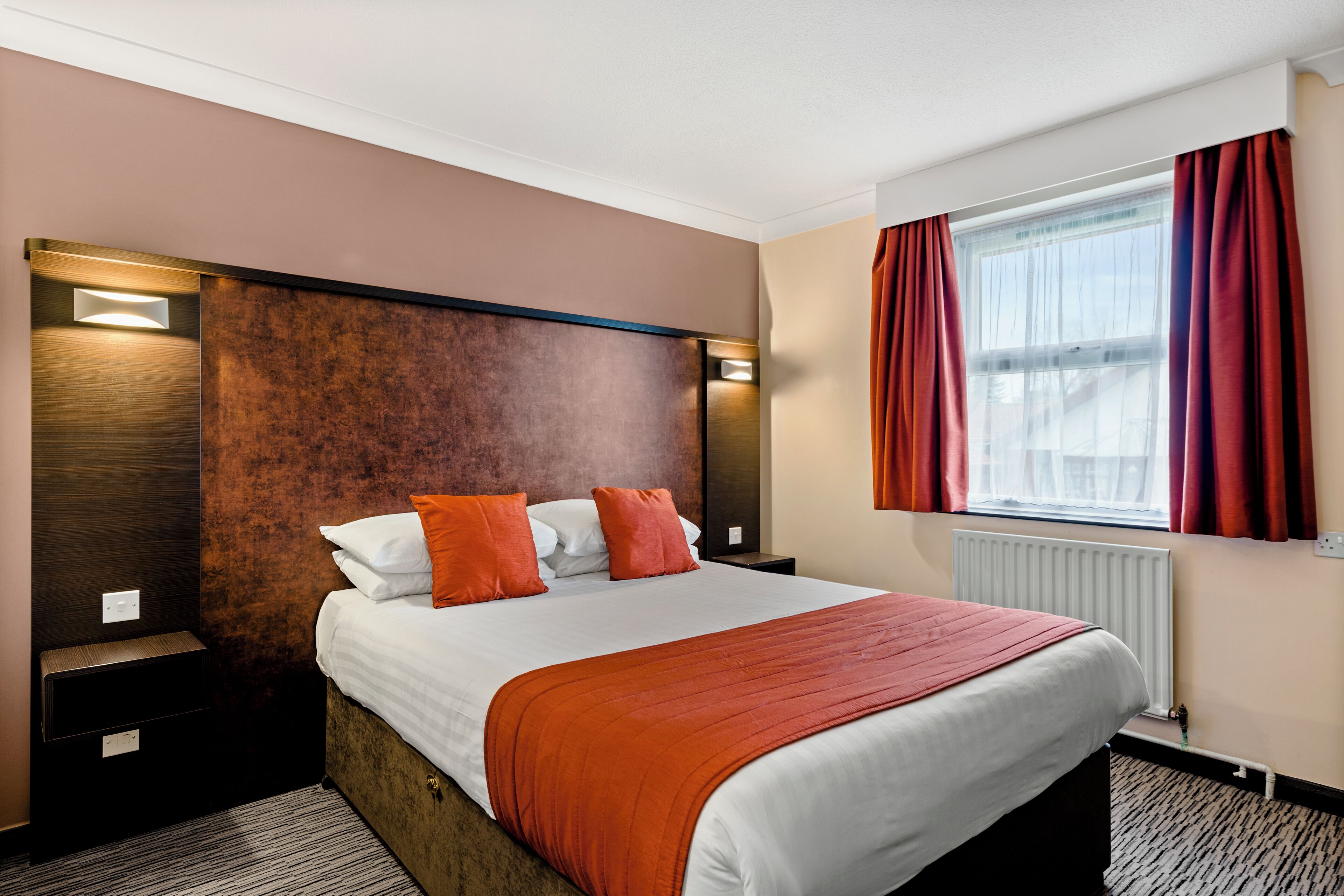 A hotel room with a large bed, a wooden headboard, a window with red curtains, and a radiator.