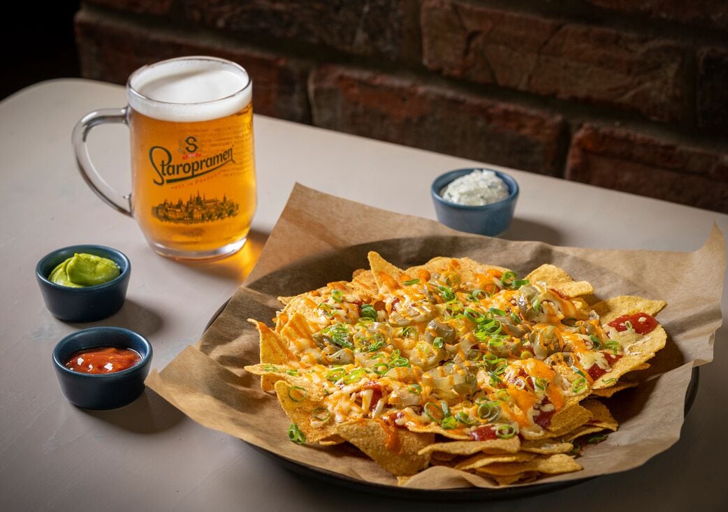 Nachos com queijo, cebolinha e jalapeños, servidos em um prato de papel com um copo de cerveja e pequenas tigelas de salsa e molho blue cheese.