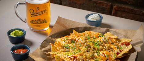 Nachos com queijo, cebolinha e jalapeños, servidos em um prato de papel com um copo de cerveja e pequenas tigelas de salsa e molho blue cheese.