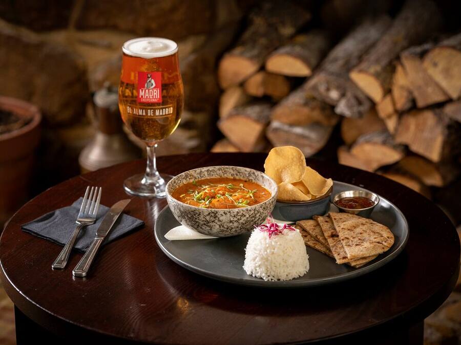 Una mesa con un tazón de sopa, arroz, pan plano, un vaso de cerveza y un recipiente pequeño de salsa.