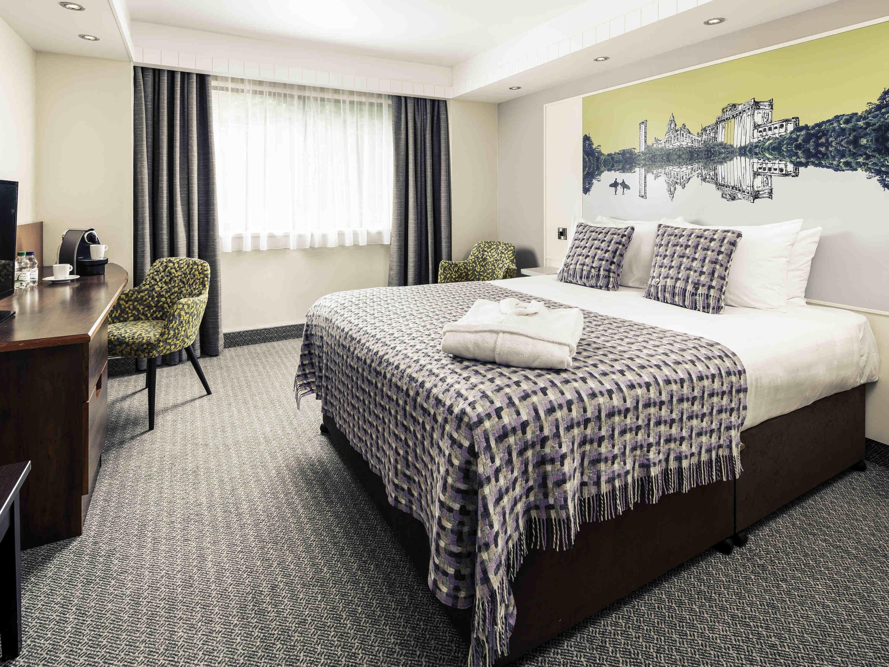 A hotel room with a large bed, a desk with a chair, a coffee machine, and a painting on the wall.
