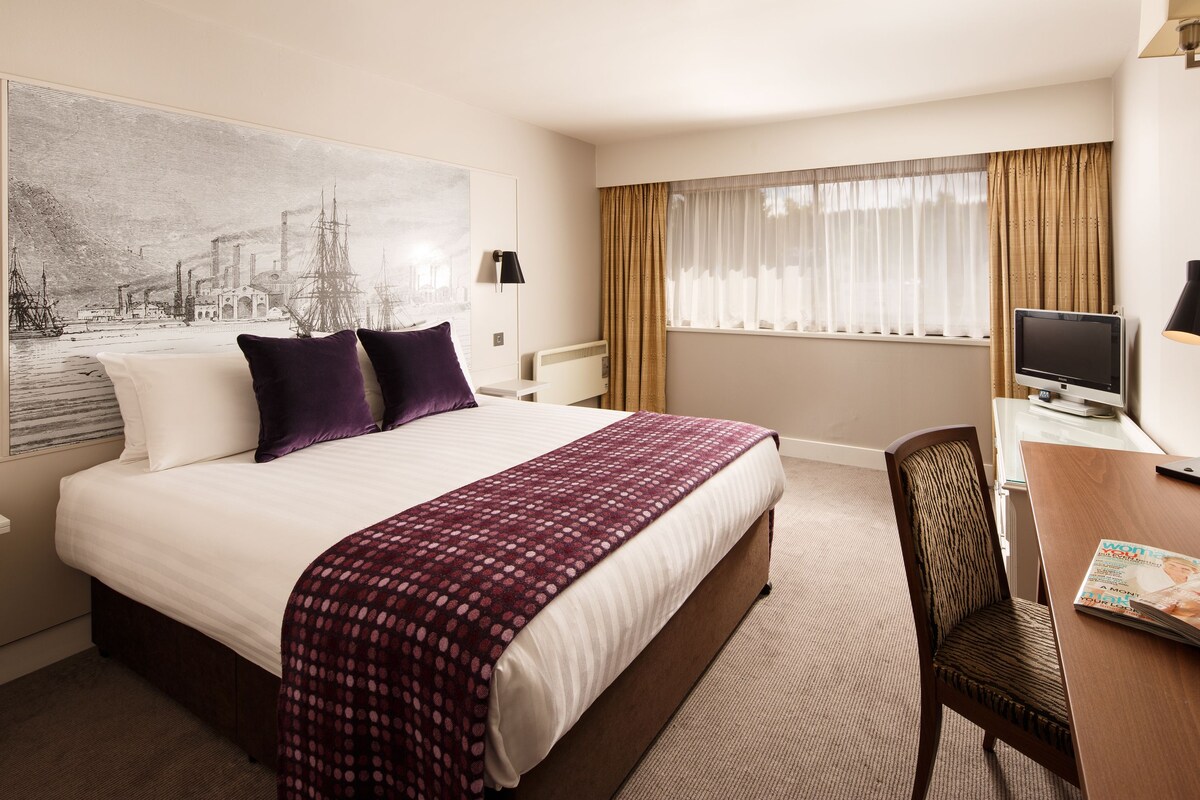 A hotel room with a large bed, a desk with a computer, a chair, and a window with curtains.