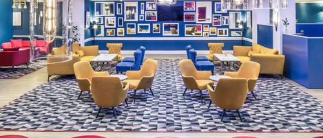 Un hall d’entrée moderne avec un sol à motifs géométriques, des espaces de détente dotés de chaises jaunes et rouges, et un mur décoré de tableaux encadrés.