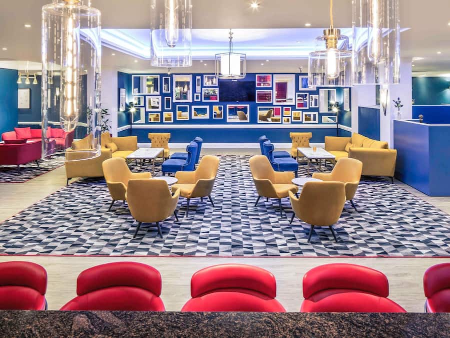 A modern lobby with a geometric patterned floor, seating areas with yellow and red chairs, and a wall decorated with framed pictures.
